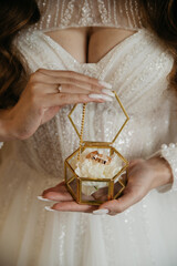 A fragile bride in a white dress with a deep neckline holds a glass box with gold wedding rings in...