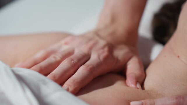 Close up of therapist's hand performing soft massage strokes on exposed collarbone and shoulder of female client lying under white towel, emphasizing relaxation, and calming therapeutic touch