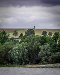 Unten das Wasser, darüber dichter Wald, dann offene Felder – und ganz oben: ein einzelner Baum unter dramatischem Himmel. Diese Szene wirkt wie ein Gemälde der Stille. Der einsame Spaziergänger.