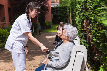 Young nurse assisting and comforting elderly women sitting on bench in the garden of a nursing home