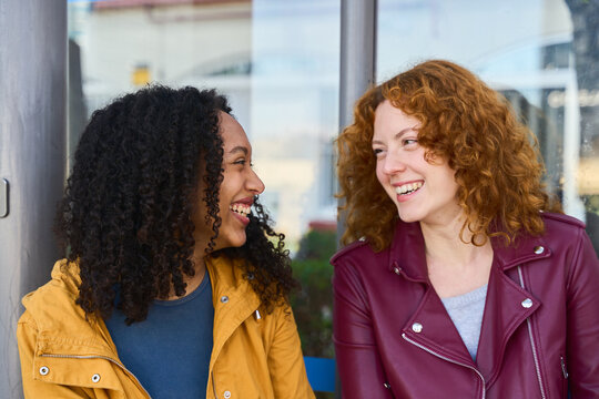 Two curly hair young women smiling and talking at the bus stop - Powered by Adobe