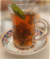 A close-up view of herbal mint tea with fresh mint leaves, presented in a clear glass on an ornate floral-patterned saucer.