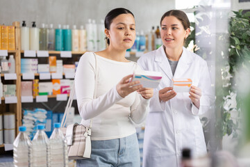 Obraz premium Young Asian woman in pharmacy holding medicine box and looking concerned while asking friendly female pharmacist for clarification before making decision