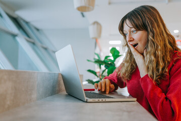 Excited businesswoman in red sweater searching online flight deals, smiling while working on laptop in contemporary workspace, celebrating affordable travel booking