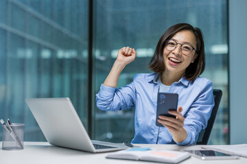 A joyful asian businesswoman celebrating a successful achievement while holding her mobile phone at...