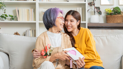 Smiling asian adult daughter greeting happy senior mother with mother's day presenting bouquet of flowers and card,happy elderly mom and grown up child hug embrace celebrate birthday at home together