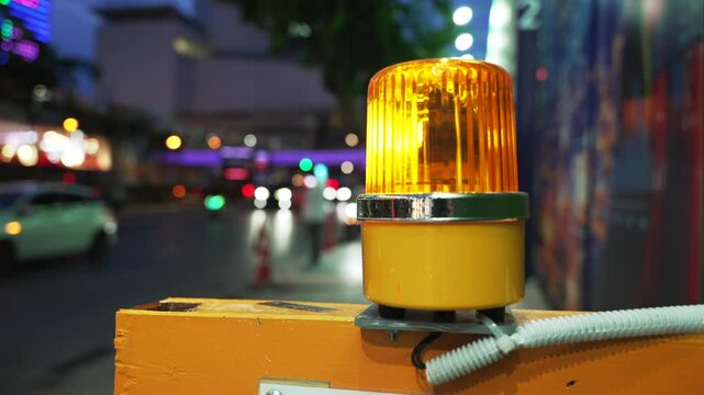 Close-up of a yellow flashing warning beacon mounted on a construction barrier at a roadside site entrance. The background is blurred with urban traffic and city lights at night during rush hour