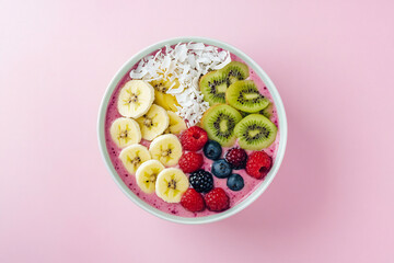 Top view of a colorful fruit smoothie bowl with banana, kiwi, berries, and coconut flakes, placed in the middle of a pastel pink background