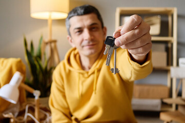 A man in a yellow hoodie holds out a set of keys towards the camera in a room with moving boxes.