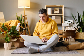 A man sits on the floor among moving boxes, using his laptop after moving into his new home.