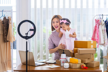 Young Asian influencer mother is selling children clothes live online streaming at home while raising her toddler as assistance