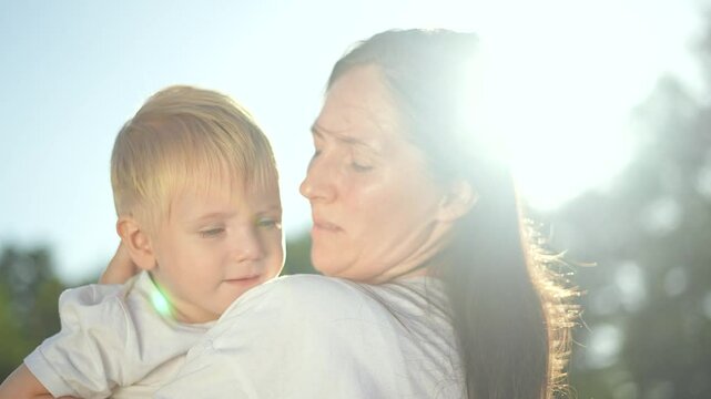 Happy Mom holding son in sunny park. Happy Tender moment of mom and son play. Loving mother and son play outdoors. Joyful park playtime between mom and son. Warm bond during playful moment in park.