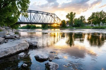 Fototapeta premium Scenic view of steel bridge over river at sunset, with reflections on water and lush greenery surrounding area