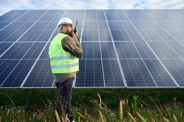 Engineer in uniform with protective helmet near solar station holding walkie talkie. Supervisor is checking on solar panels with radio station on solar farm. Solar and renewable energy concept.