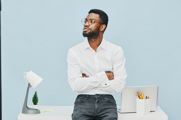 Confident young man with glasses, black hair, and casual white shirt standing with crossed arms behind a desk in a modern office with a minimalist light blue background, creating a professional