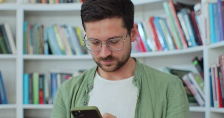 Portrait of happy young student chatting by mobile phone in university library. Young man wearing green shirt and eyeglasses using social media application browsing on smartphone. University library - Powered by Adobe