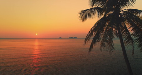 Orange and red sunset over calm sea water with silhouette of palm tree leaves in the foreground and small islands in the distance, Koh Samui, Thailand