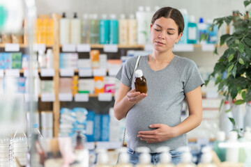 Young pregnant woman buyer choosing medicinal cough liquid syrup standing between shelves at small...