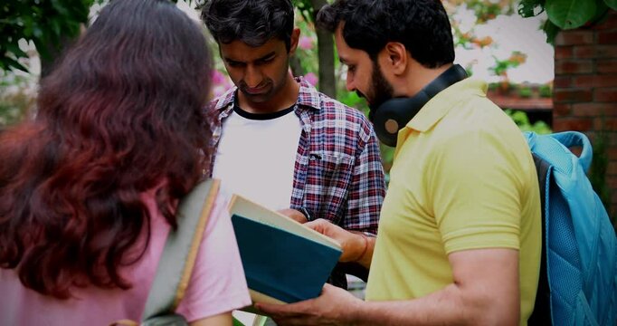 Standing students discussing project outdoors as Indian university classmates have a chat with laptop, book, and notes in college campus garden, sharing ideas and fruitful conversation together