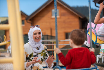 A happy Muslim family is having fun in the park, a young smiling mother in hijab pushes her child on a swing in the garden playground.