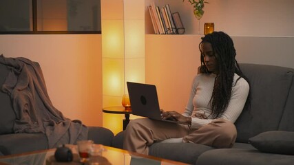 Young African American woman with dreadlocks sitting on sofa at home in the evening, calmly working on laptop with thoughtful expression and gentle smile in a cozy, relaxed setting. - Powered by Adobe