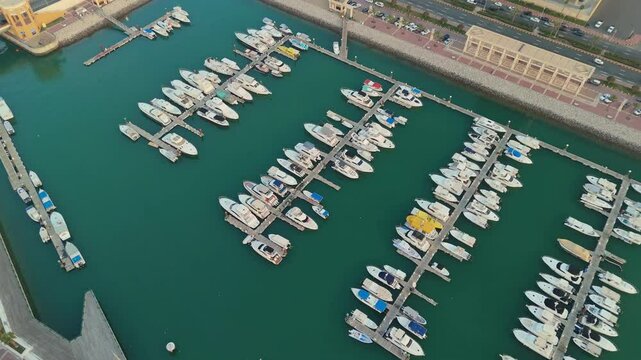 High-resolution 4K drone view of a luxury marina in Kuwait, showing boats lined up along docks with clear blue waters and breakwater paths. Perfect for travel and architecture visuals.