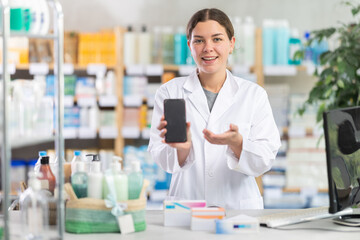 Friendly skilled young girl pharmacist standing behind counter, demonstrating and offering mobile application for smartphone in modern pharmacy