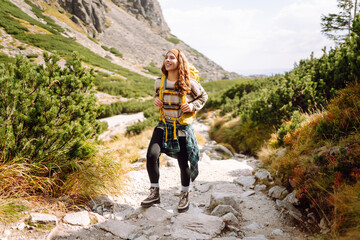 Young female hiker with yellow hiking backpack on rocky trail with mountain view. Happy woman enjoying weather and outdoor adventure. Travel concept.