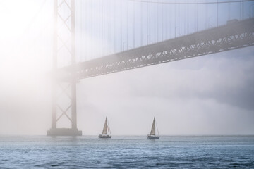 25 de Abril bridge on foggy sunny day with sailboats on Tagus river