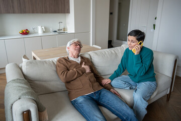 Concerned wife makes urgent call during husband heart attack. Senior male collapsing, chest pain, panic in room, aging health risk, cardiovascular failure, immediate assistance and domestic emergency