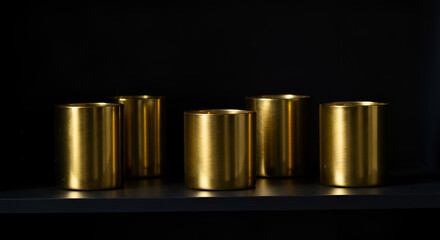 Gold candle holders on a matte black shelf, dim ambient glow revealing gentle contours and subtle shimmer in a shadowed space.