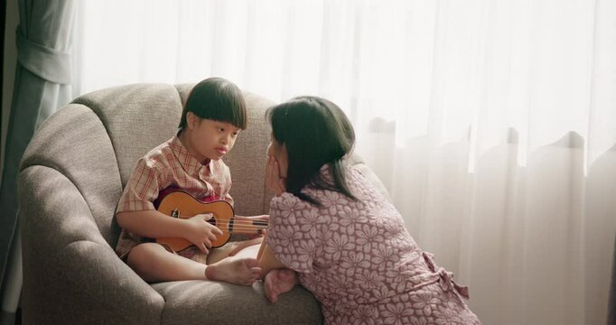 Asian young boy with Down syndrome holding ukulele and singing near window with natural light. Supportive mother listening. Emotional connection, music therapy, special needs care.