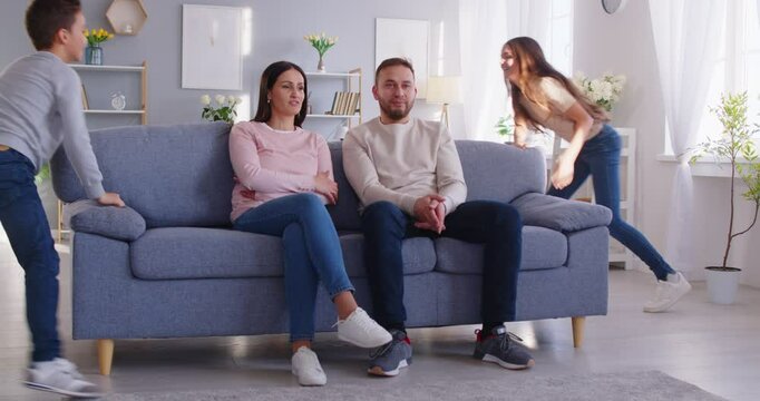 Exhausted and tired parents sitting on the couch while kids playing, run around, and create noise at home. Misbehave, chaos, stress, and challenges of parenting, balanced with moments of family life.