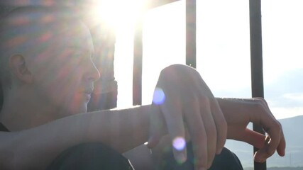 A shaved-head teenage boy sits quietly by a barred prison window, staring out with a sad, thoughtful gaze. A young inmate reflecting on his past, regretful and hoping for a second chance.