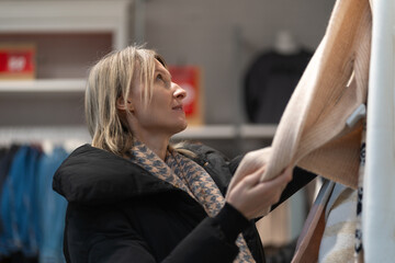 Blonde woman wearing a stylish black jacket is shopping in a clothing store, reaching out to grab an item from a rack surrounded by a softly blurred background