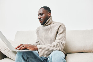 Young African man working on a laptop at home, wearing a stylish beige sweater and relaxed jeans, exuding a focused and content emotion in a cozy environment