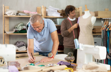 Fototapeta premium Senior man tailor draw dress pattern of product on paper, other work with mannequin. Craftsman perform paper prototype of product parts, drawing paper pattern for dressmaking.