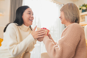 A loving asian daughter gives her mother a birthday gift while sitting together on the couch,...