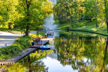 A clean and calm Riga city canal surrounded by trees with spring foliage and a wooden pier for pleasure boats, Riga. Latvia.