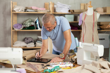 Elderly male tailor draws pattern using tablet in sewing workshop
