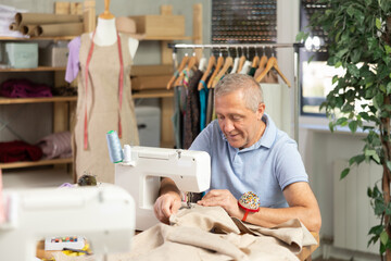 Senior man tailor at work. Master sews clothes in sewing machine. Worker uses sewing machine to...