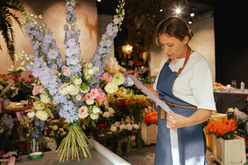 Middle-aged female florist in apron arranging fresh flowers for bouquet She ties a ribbon on the...