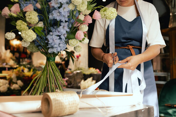 Middle-aged female florist in apron arranging fresh flowers for bouquet She ties a ribbon on the...