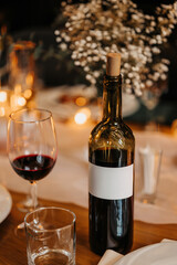 Vertical photo with a wine glass, in the background you can see a beautiful table setting. A bottle of wine rests on a restaurant table, surrounded by candlelight—warm, romantic, and inviting.
