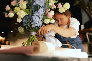 Middle-aged female florist in apron arranging fresh flowers for bouquet She ties a ribbon on the...