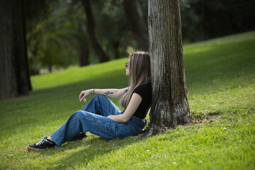Obraz premium Young, beautiful blonde woman, wearing a black t-shirt, jeans, and sunglasses, relaxed, calm, and solitary, sitting on the grass leaning against a tree resting in a park. Concept of solitude, peace.