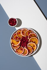 Rodajas de naranja sanguina y semillas de granada en un plato blanco sobre un fondo blanco y azul....