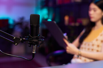 Woman working on podcast content plan, while preparing for livestreaming, with professional microphone in studio. Colorful lighting creating artistic ambiance