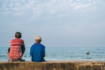 backs of two sad men elderly retired fishermen sitting on beach by the sea