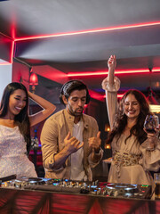 Group of stylish young adults enjoying a party in a nightclub with red neon lights. A DJ is mixing music while two women cheer with glasses of red wine, smiling and having fun.